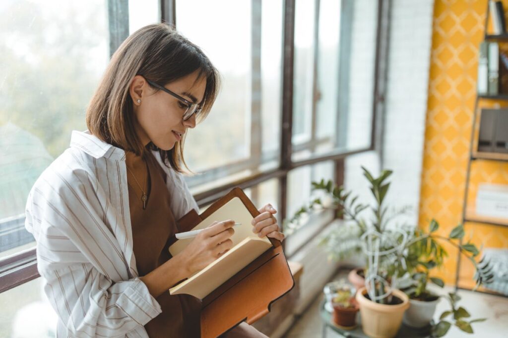 Eine Frau, die am Fenster lehnt und in ein Tagebuch schreibt, symbolisiert den Prozess der inneren Einkehr und des Erkennens von inneren Blockaden