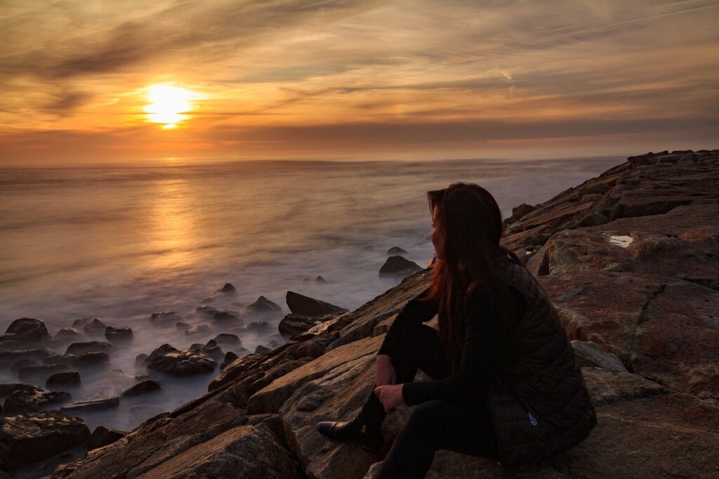 Eine Frau sitzt auf einem Hügel am Meer und beobachtet den Sonnenuntergang, symbolisch für die innere Ruhe und Reflexion nach der Anwendung der Yager Methode.